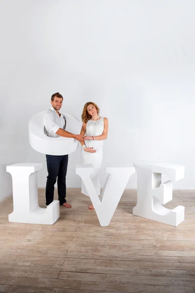 Gelukkige zwangere paar gekleed in het wit op witte achtergrond met gigantische handgemaakte letters van het woord liefde — Stockfoto