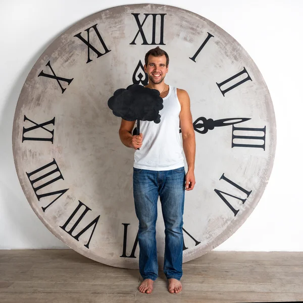 Felice uomo sorridente vestito di bianco in piedi scalzi sul pavimento di legno e mostrando striscione segno discorso bolla cercando eccitato e avere idea su sfondo bianco con orologio gigante . — Foto Stock