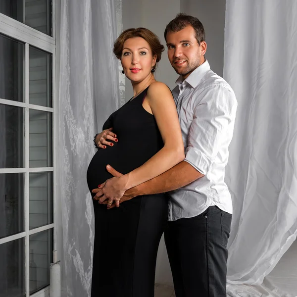 Feliz casal grávida vestido de preto e branco abraçar uns aos outros pela janela — Fotografia de Stock