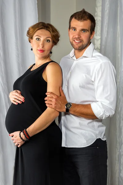 Happy Pregnant Couple dressed in black and white embrace each other by the window — Stock Photo, Image