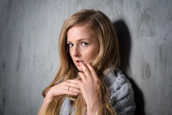 Sexy joven mujer rubia de pelo largo en suéter de punto posando contra la pared gris gruesa. Chica atractiva asustada o resentida escondida detrás de un mechón de su cabello —  Fotos de Stock