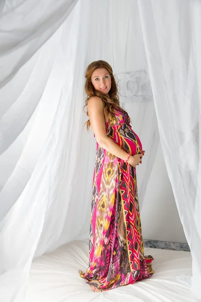 Young beautiful fashion pregnant woman in colorful dress standing on a bed with a canopy on a white background — Stok fotoğraf