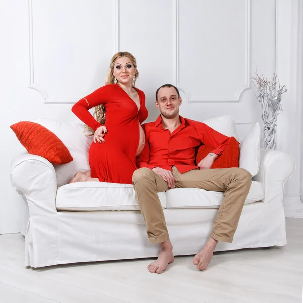 Happy young pregnant couple dressed in red embrace each other on a sofa in white living room — Stock Photo, Image