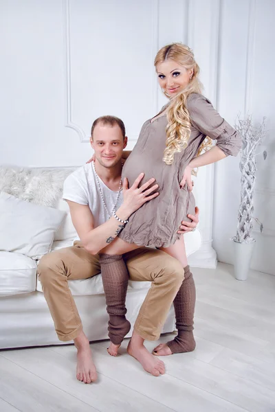 Happy young pregnant couple embrace each other on a sofa in white living room — Stock Photo, Image