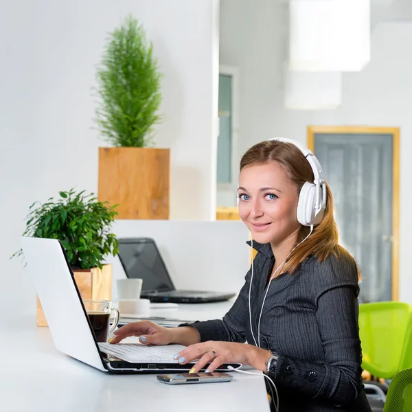 Νέοι επιτυχημένη επιχειρηματίας ακουστικά με φορητό υπολογιστή, σερφάρισμα στο Διαδίκτυο στο σύγχρονο γραφείο φωτεινά. Γυναίκα χρησιμοποιώντας υπολογιστή δισκίο και να ακούτε τη μουσική κατά τη διάρκεια της διάλειμμα για καφέ. — Φωτογραφία Αρχείου