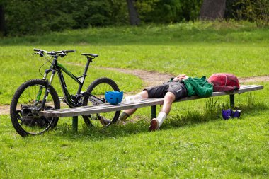 Cyclist resting on a bench in the park. Tired young man with his mountain bike in the forest resting and making pause.