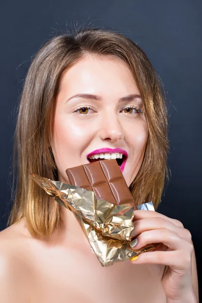 Beautiful  naked young blonde woman with bright makeup eating chocolate bar. — Stockfoto