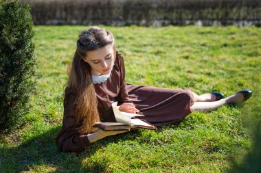 Bahçede kitap okuyan romantik genç bir kadın çimlerin üzerinde oturuyor. Açık hava zaman kavramını gevşetin.