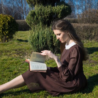 Bahçede kitap okuyan romantik genç bir kadın çimlerin üzerinde oturuyor. Açık hava zaman kavramını gevşetin.