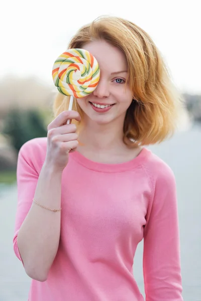Redhead beautiful young woman holding a lollipop in front of her eye. Pretty girl having fun outdoors. — Stok fotoğraf