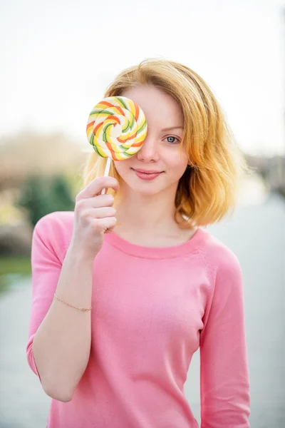 Redhead beautiful young woman holding a lollipop in front of her eye. Pretty girl having fun outdoors. — Stok fotoğraf