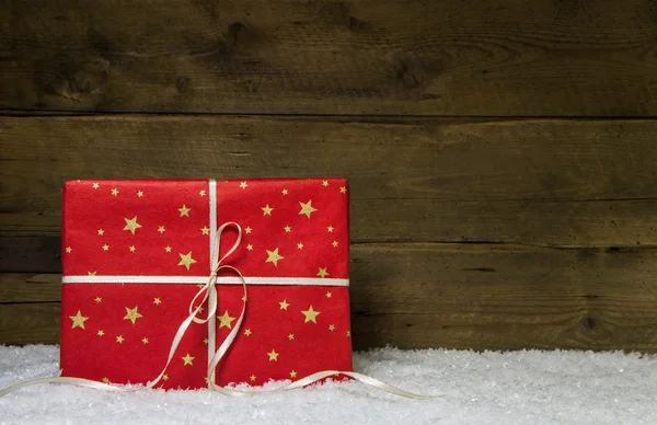 Un regalo de Navidad rojo con estrellas doradas en la espalda nevada de madera —  Fotos de Stock