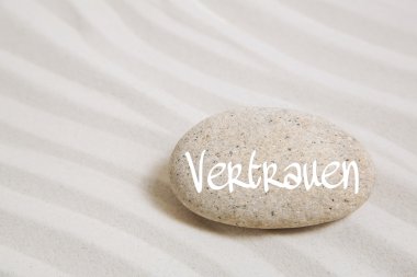 Sand background with a stone and the german word for trust. clipart