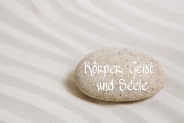 Stone in the sand with the german text: body, mind and soul. Bac clipart