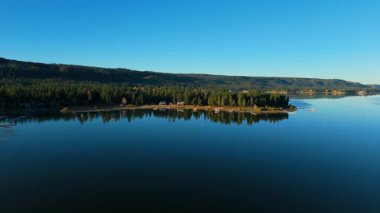 İnsansız hava aracı görüntüleri Güney Kaliforniya 'daki Big Bear Gölü' nün geniş bir görüntüsünü gösteriyor. Sakin yansıtıcı su, ormanlık kıyı şeridi ve dağınık göl evleri var. Erken sabah ışığı sonbaharı vurgular