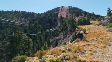 Kaliforniya, Big Bear Gölü 'nde açık bir sonbahar gününde orman yamacının üzerinde bir dağ kaldıracı boyunca kayar. Renkli sonbahar çalıları ve çam ağaçları, parlak mavi bir gökyüzünün altındaki yamacı kaplar.