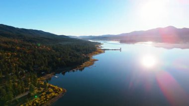 Sinema insansız hava aracı günbatımında Büyük Ayı Gölü 'nü sakin yansıtıcı su ve yoğun yeşil ormanlarla çevrili uzun bir kıyı şeridiyle yakalar. Altın sonbahar ağaçları ve yumuşak akşam ışığı