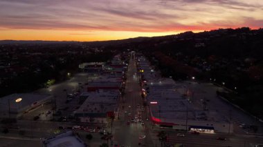 Akşam trafiği, aydınlatılmış mağazalar ve renkli günbatımı ve erken gece geçişi sırasında çevreleyen tepelerin bulunduğu ana şehir caddesinin insansız hava aracı görüntüsü. 28 Kasım 2025 'te çekildi. Stüdyo Şehri