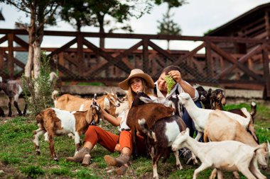 Mutlu bir aile, şapkalı bir adam ve bir kadın, güneşli bir yaz gününde, keçilerle çevrili bir çiftlikte rahatlıyor ve gülüyorlar.