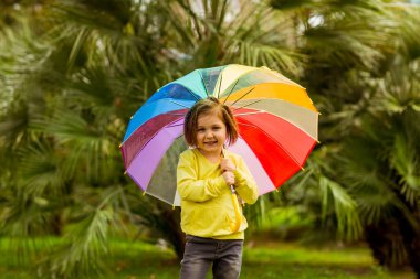 Sarı kazak, kot pantolon ve sarı çizme giyen güzel küçük kız yeşil palmiye ağaçlarının arka planında renkli bir şemsiyeyle dikiliyor.