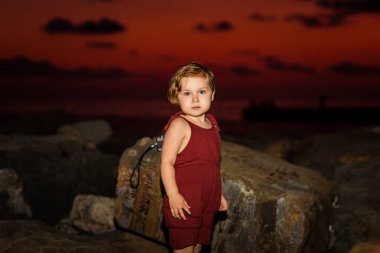 Küçük güzel bir kız, kumsalda, bir yaz akşamı, güzel bir gün batımı ve bulutlarla, elinde bir kamerayla oturuyor.