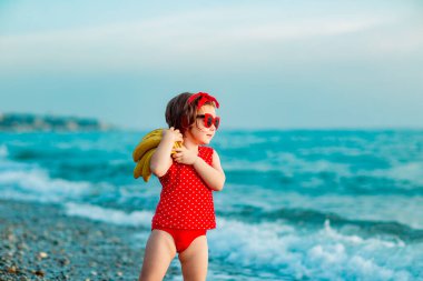 Kırmızı mayolu ve güneş gözlüklü küçük bir kız sahilde çıplak ayakla duruyor ve olgun muzlar tutuyor.