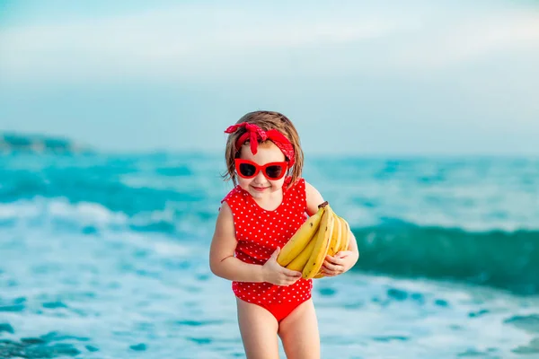 Little Girl Red Swimsuit Sunglasses Standing Barefoot Beach Holding Ripe — Stock Photo, Image