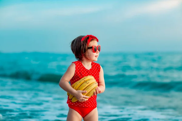 Little Girl Red Swimsuit Sunglasses Standing Barefoot Beach Holding Ripe — Stock Photo, Image