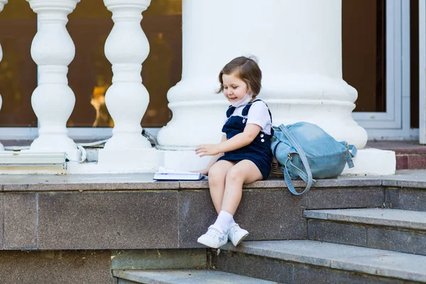 学校の制服を着た女の子と医療用のマスク学校の建物の近くのリュックと日記に座って — ストック写真