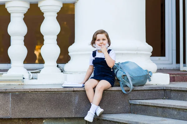 学校の制服を着た女の子と医療用のマスク学校の建物の近くのリュックと日記に座って — ストック写真