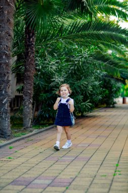 a little girl, a schoolgirl, goes to school in the park with a denim backpack, and in a medical mask