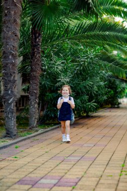 a little girl, a schoolgirl, goes to school in the park with a denim backpack, and in a medical mask