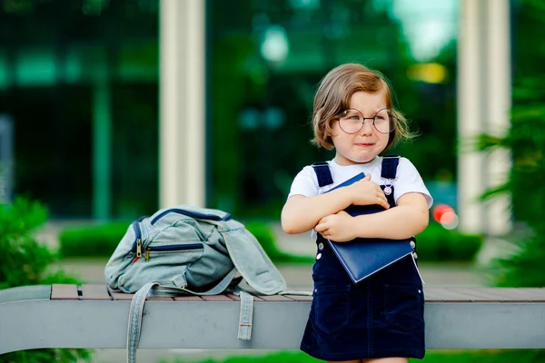 小さな女の子が学校の近くに立っていて制服と眼鏡を着ていて手に日記をつけて悲しい気分で泣いています — ストック写真