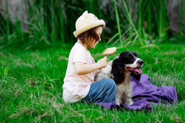 Şapkalı küçük, güzel bir kız, en sevdiği köpeği tarıyor, parkta battaniye ve yeşil çimenler üzerinde.