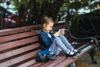 Okul üniformalı küçük, güzel bir kız bankta oturuyor ve elinde bir tablet tutuyor.
