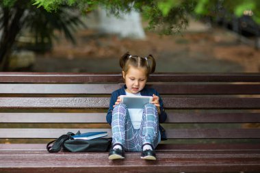Okul üniformalı küçük, güzel bir kız bankta oturuyor ve elinde bir tablet tutuyor.