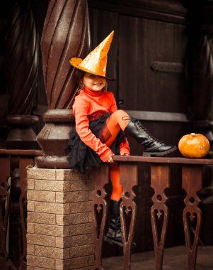 Küçük, güzel bir kız, turuncu ve siyah bir cadı kostümü içinde, Cadılar Bayramı 'nda, tahta bir evin verandasında oynar.