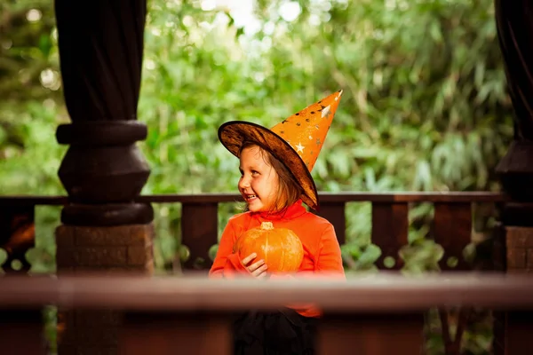 万圣节那天 一个身穿橙色和黑色女巫服装 头戴南瓜的漂亮小女孩在一座木头房子的门廊上玩耍 — 图库照片
