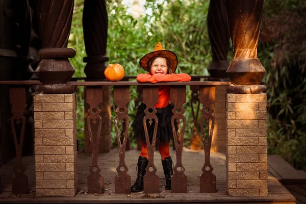 万圣节那天 一个身穿橙色和黑色女巫服装 头戴南瓜的漂亮小女孩在一座木头房子的门廊上玩耍 — 图库照片