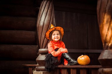 Küçük, güzel bir kız, turuncu ve siyah bir cadı kostümü içinde, Cadılar Bayramı 'nda, tahta bir evin verandasında oynar.