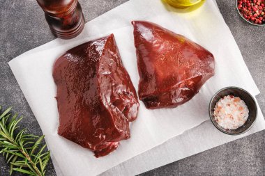 Raw cow liver on white paper with salt, pepper and green,  top view, high angle