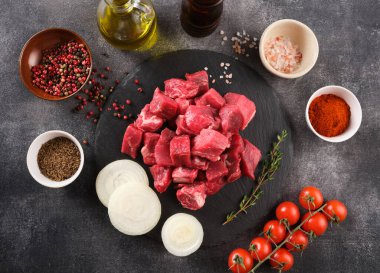 Fresh raw chopped beef on an stone board, spices, herbs and vegetables on dark grey background. Raw ingredients for goulash