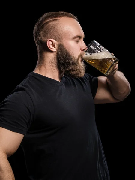 Hombre barbudo bebiendo cerveza de una taza de cerveza sobre fondo negro . — Foto de Stock