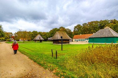 Kırsal kesimde küçük ilkel kulübeler, toprak yolda yürüyen kadınlar, arka planda çiftlik evleri ve yapraklı ağaçlar, sazlı ve kiremitli çatılar, Bokrijk Açık Hava Müzesi 'nde bulutlu bir gün, Genk, Belçika