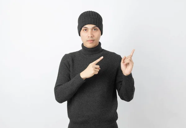 若いですハンサムなアジアの男を着てグレーのセーターとビーニーを指差す側に指で製品やアイデアを提示する白い背景に — ストック写真