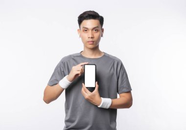Spor kıyafetleri içindeki yakışıklı Asyalı genç adam boş ekranlı telefonu gösteriyor ve beyaz arka planda izole edilmiş müzik dinliyor..