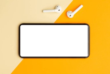 Düz yerde, turuncu ofis masasının üst görüntüsü akıllı telefon boş ekran maketi kırpma yolu ve kablosuz kulaklıkları var. Modern çalışma alanı.