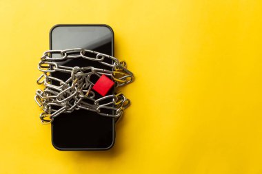Zincire vurulmuş bir akıllı telefonun yüksek açısı ve sarı arka planda kırmızı bir asma kilit. Dijital detoks kavramı, sosyal medya bağımlılığı, akıllı telefon kısıtlaması ve zihinsel sağlık.