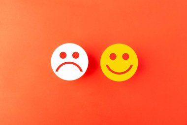 Beyaz üzgün yüz ve sarı gülen yüz simgeleri turuncu arka planda bir araya getirilmiş. Müşteri geribildirim, memnuniyet derecesi, duygusal karşılaştırma ve iş değerlendirmesi kavramı.