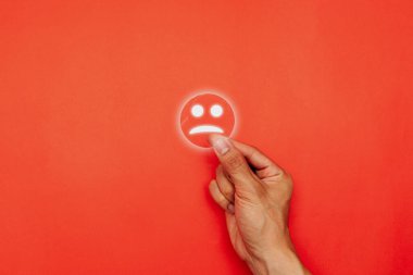 Kırmızı arka planda hüzünlü bir yüz simgesini tutan el. Kötü müşteri deneyimi, olumsuz geri bildirim, zihinsel sağlık stresi ve hizmet iyileştirme veya destek gerektiren iş sorunları kavramı.